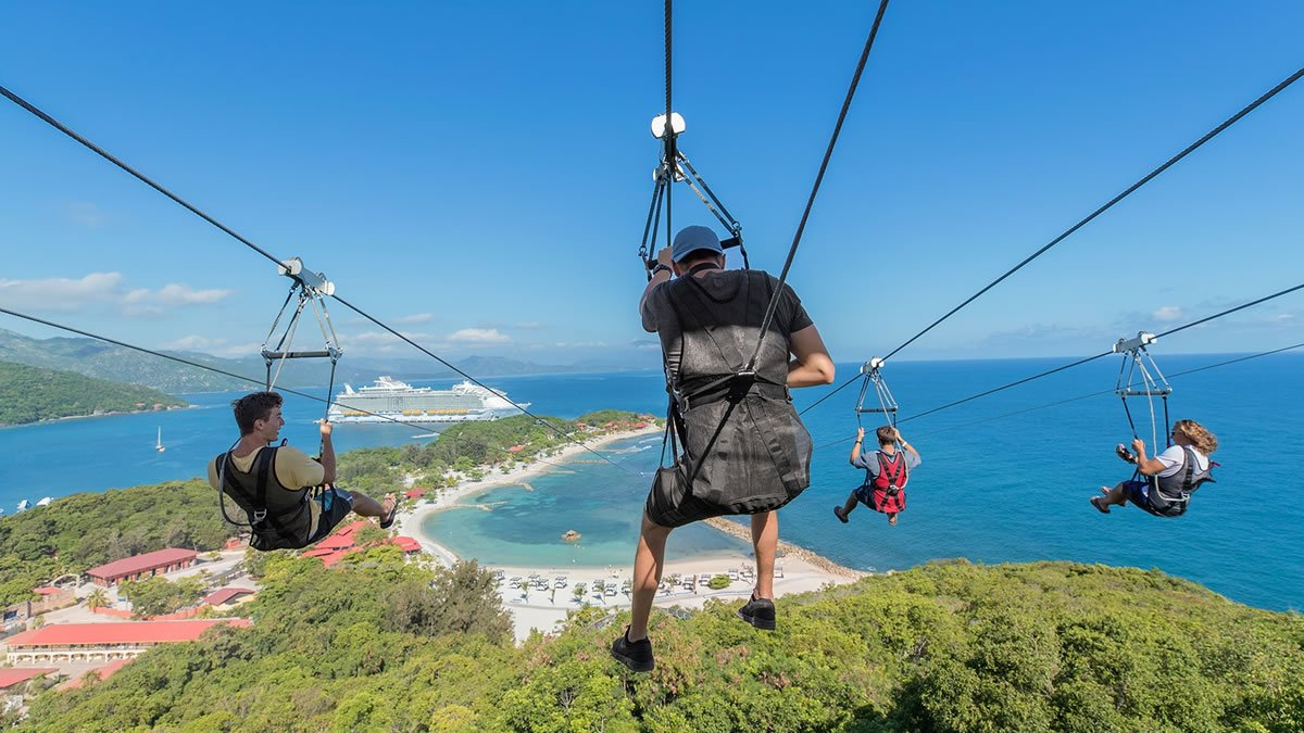 People riding a high-speed zip line over lush green hills and ocean bay with a cruise ship anchored in the distance.