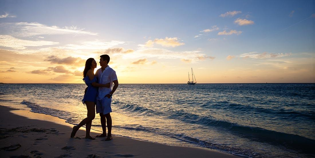 Couple embracing on a sandy Caribbean beach at sunset with a sailboat anchored on the horizon.