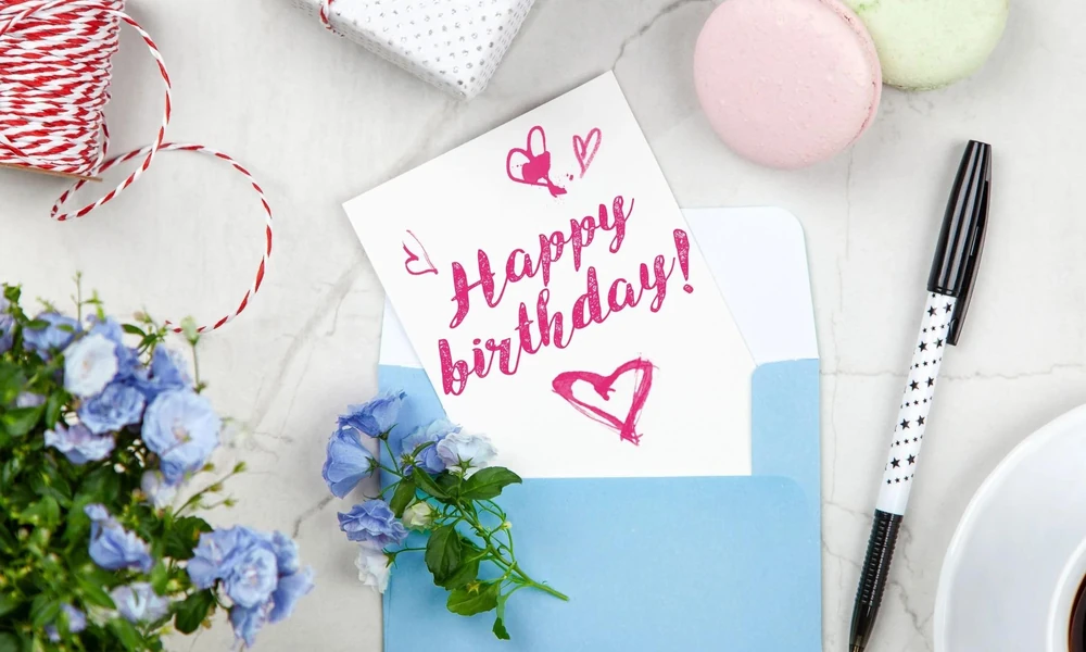 A handwritten Happy Birthday card inside a blue envelope next to flowers and a pen.