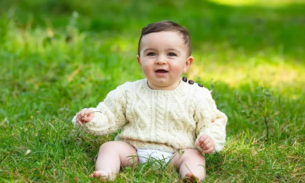 A happy baby boy sitting on grass wearing a traditional cream Aran cable-knit sweater.