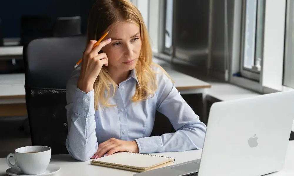 A frustrated woman sitting at a desk with a laptop and a cup of coffee, holding a pencil to her head-Funny Puns