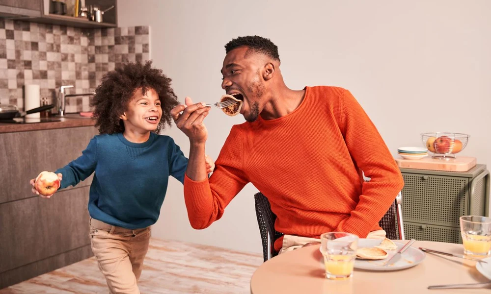 A happy father and child laughing while eating breakfast together in the kitchen.