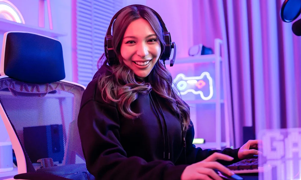 A smiling woman at her PC with a gaming headset under purple ambient lighting, searching for girl gamer nicknames