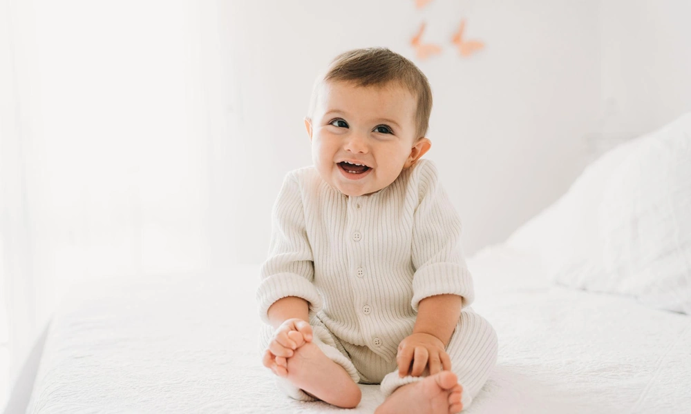 Happy baby wearing a cream ribbed knit romper sitting on a white bed and smiling.