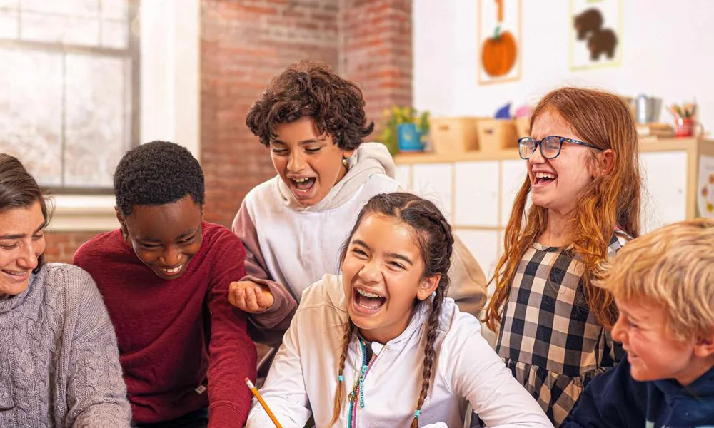 A group of diverse children and a teacher laughing together around a table.