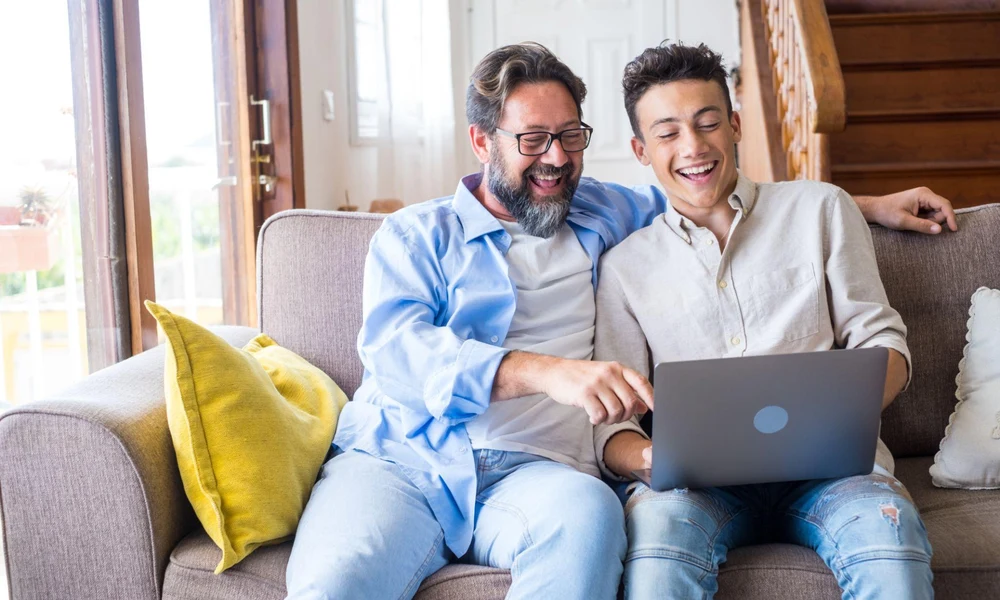 A father and teenage son laughing together while looking at a laptop on a living room couch.