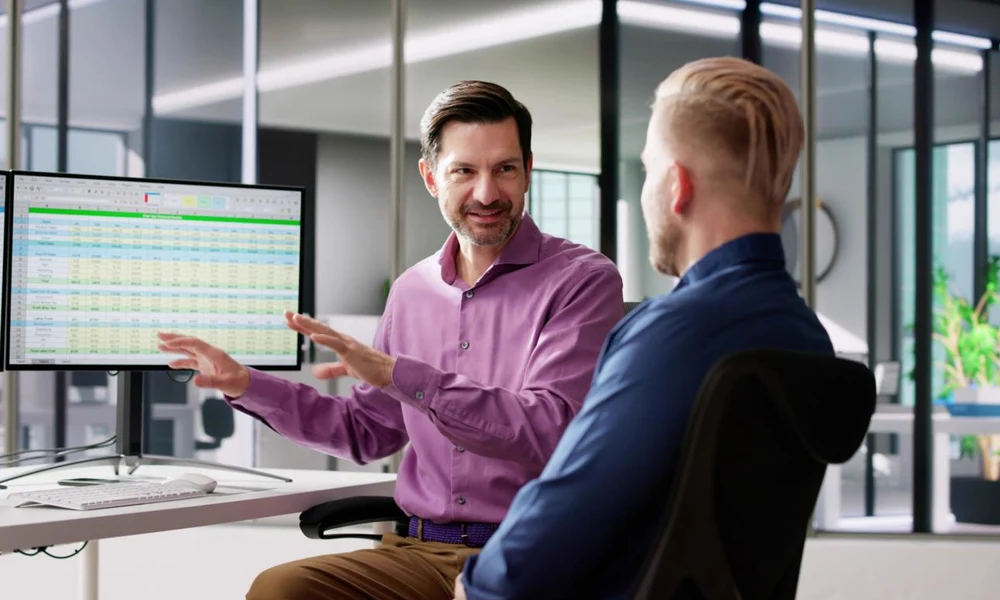 Two office colleagues discussing a spreadsheet on a computer monitor in a modern workspace-Best Dad Jokes and Puns